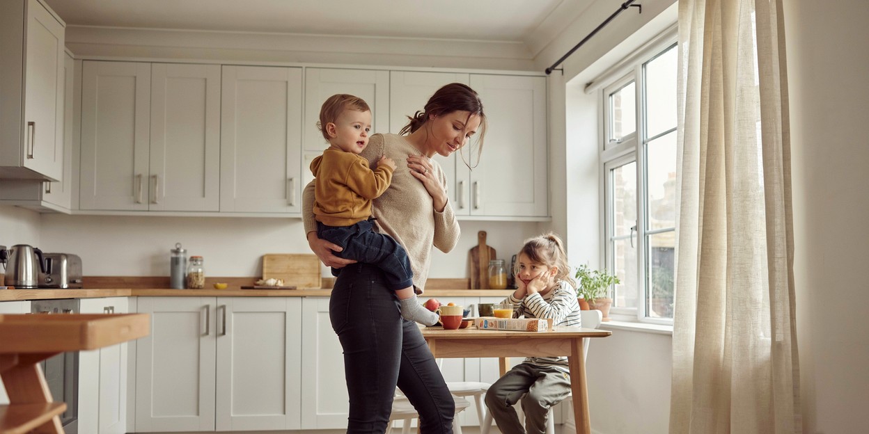 Woman in her thirties holding her toddler on one hip in a bright kitchen while an older child sits at the table